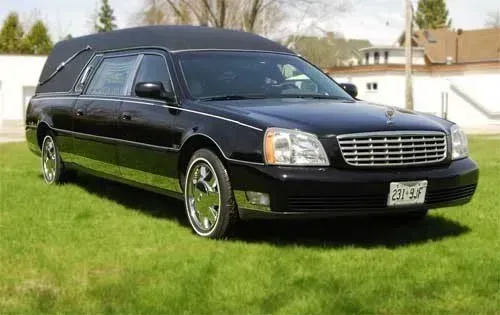 Black hearse parked on a grassy lawn in front of houses, viewed from the front-left side