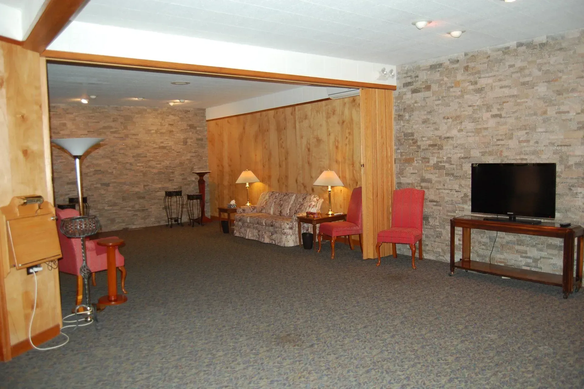 Spacious basement living room with carpet, stone walls, sofa, lamps, red chair, and TV on a stand
