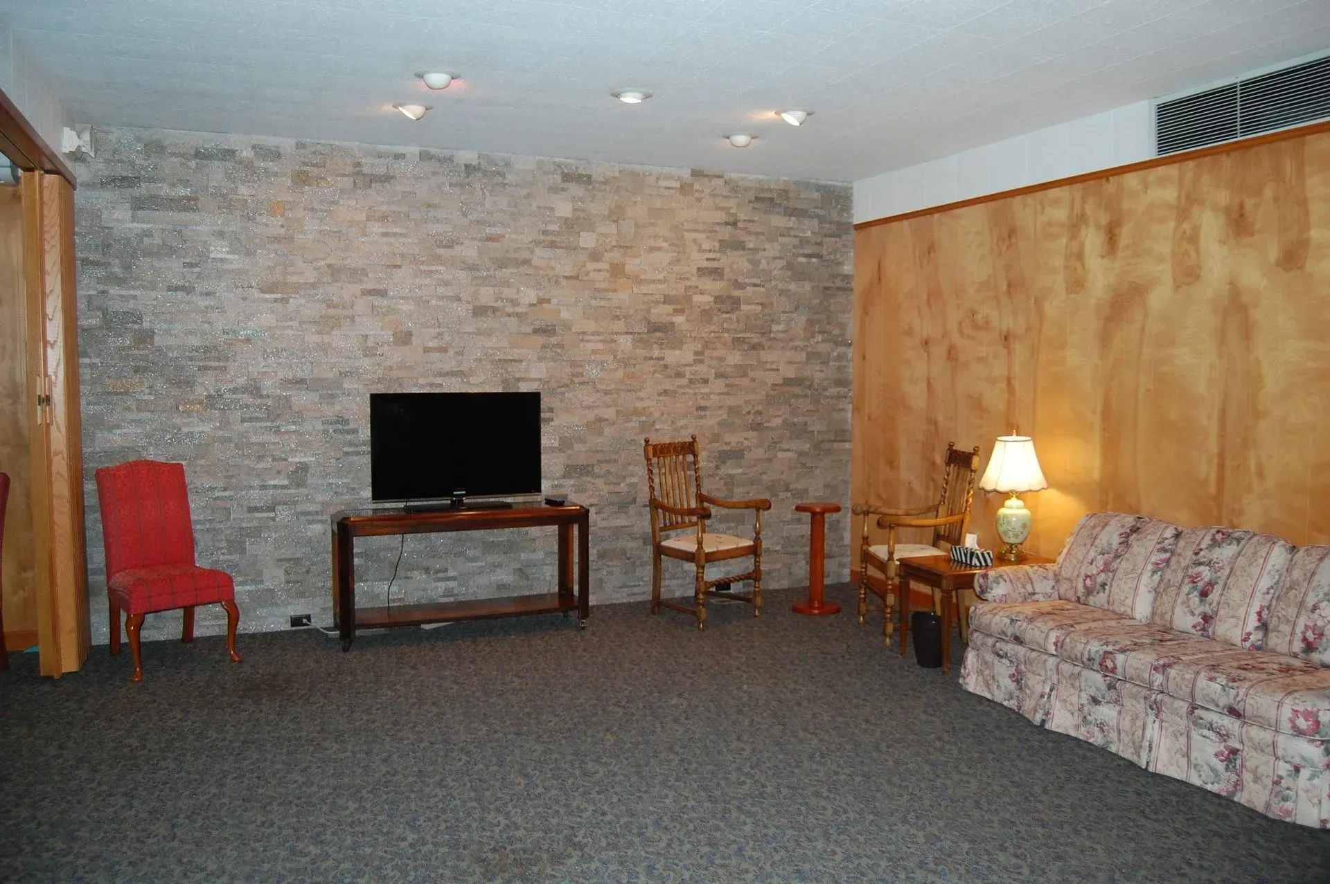 Living room with stone accent wall, TV, patterned sofa, lamp, and two chairs on carpet.