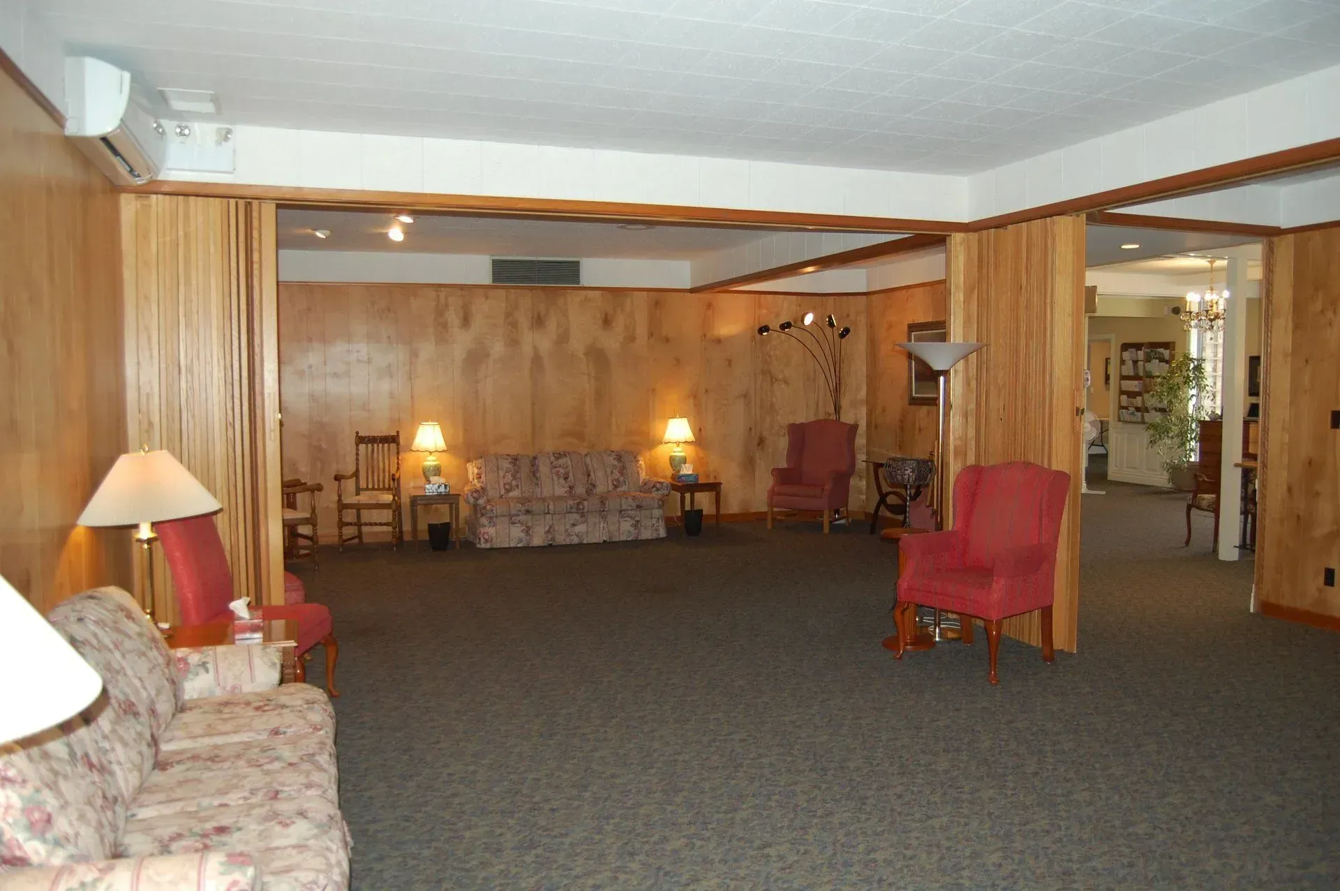 Empty carpeted lounge with wood-paneled walls, red chairs, and warm lamp lighting