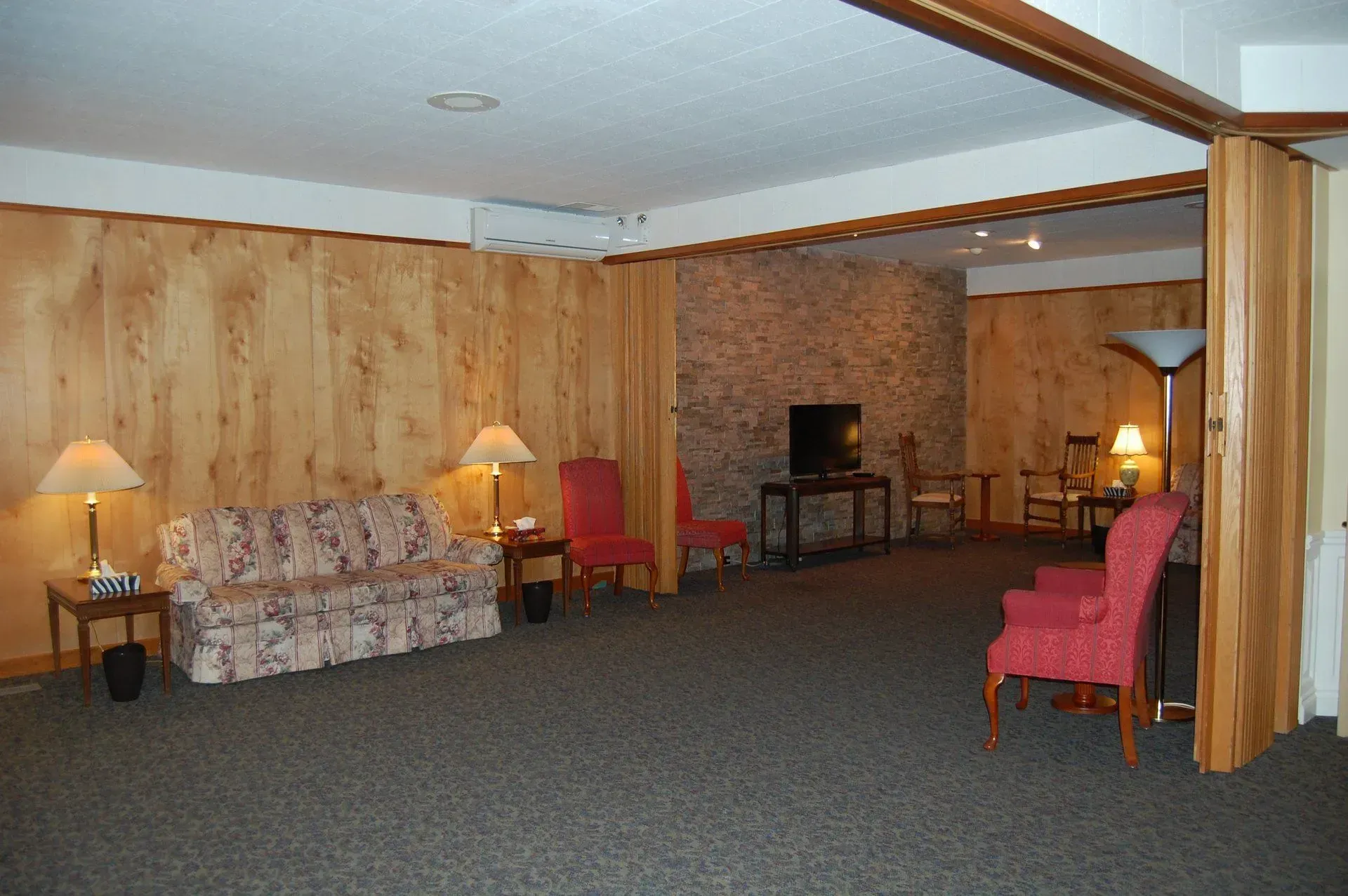 Wood-paneled hotel lounge with floral sofa, red chairs, lamps, and a small TV on a table
