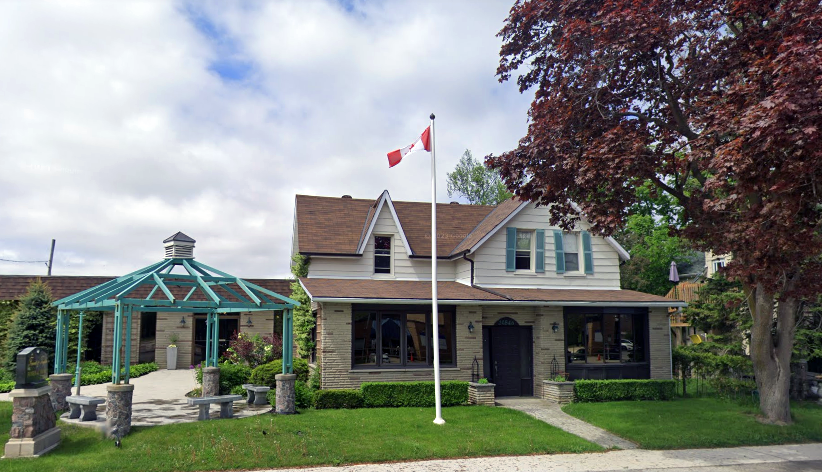 Historic stone house with green gazebo, white flagpole, and Canadian flags by a tree-lined street