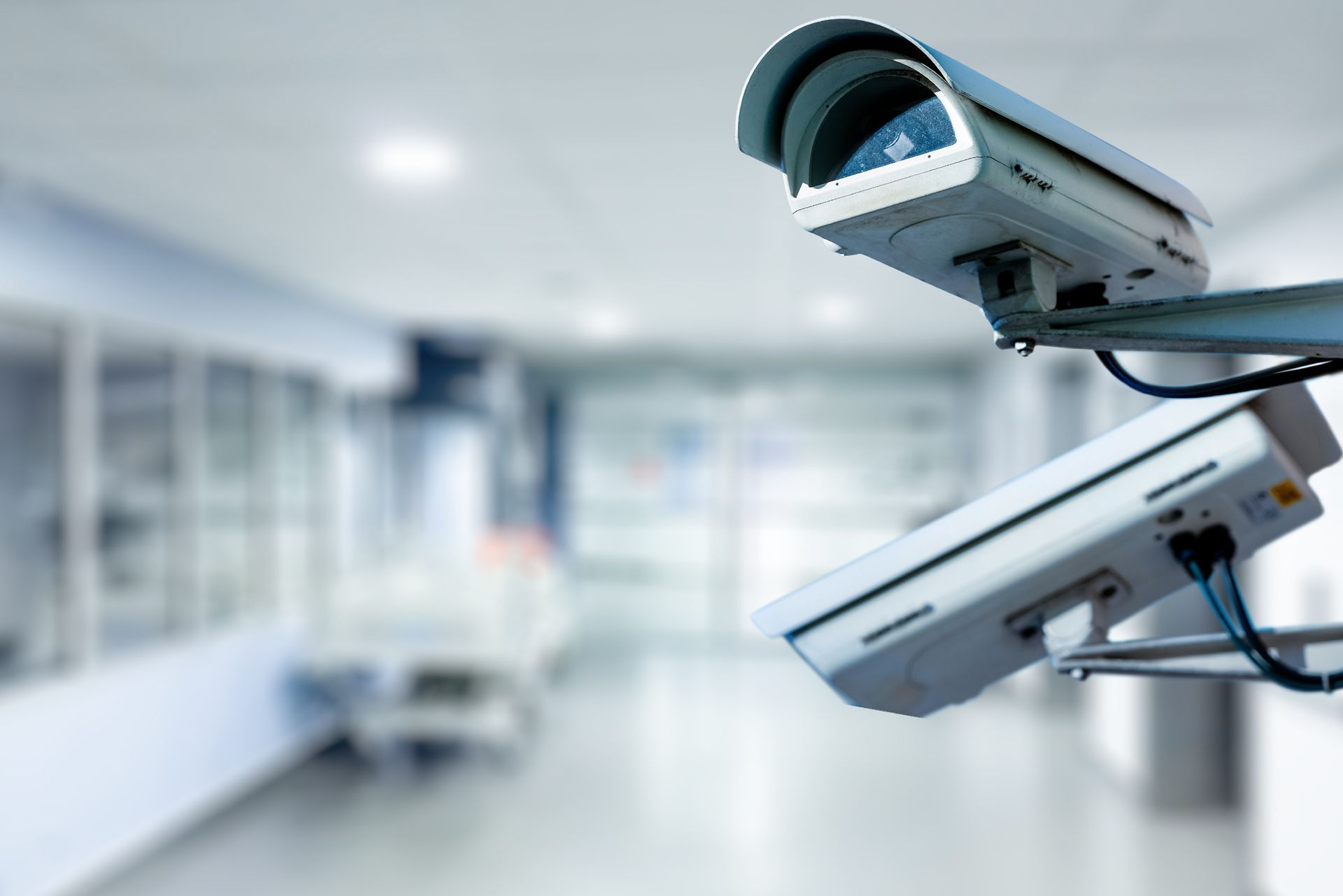Two security cameras, white, mounted, overlooking a blurred hospital hallway.
