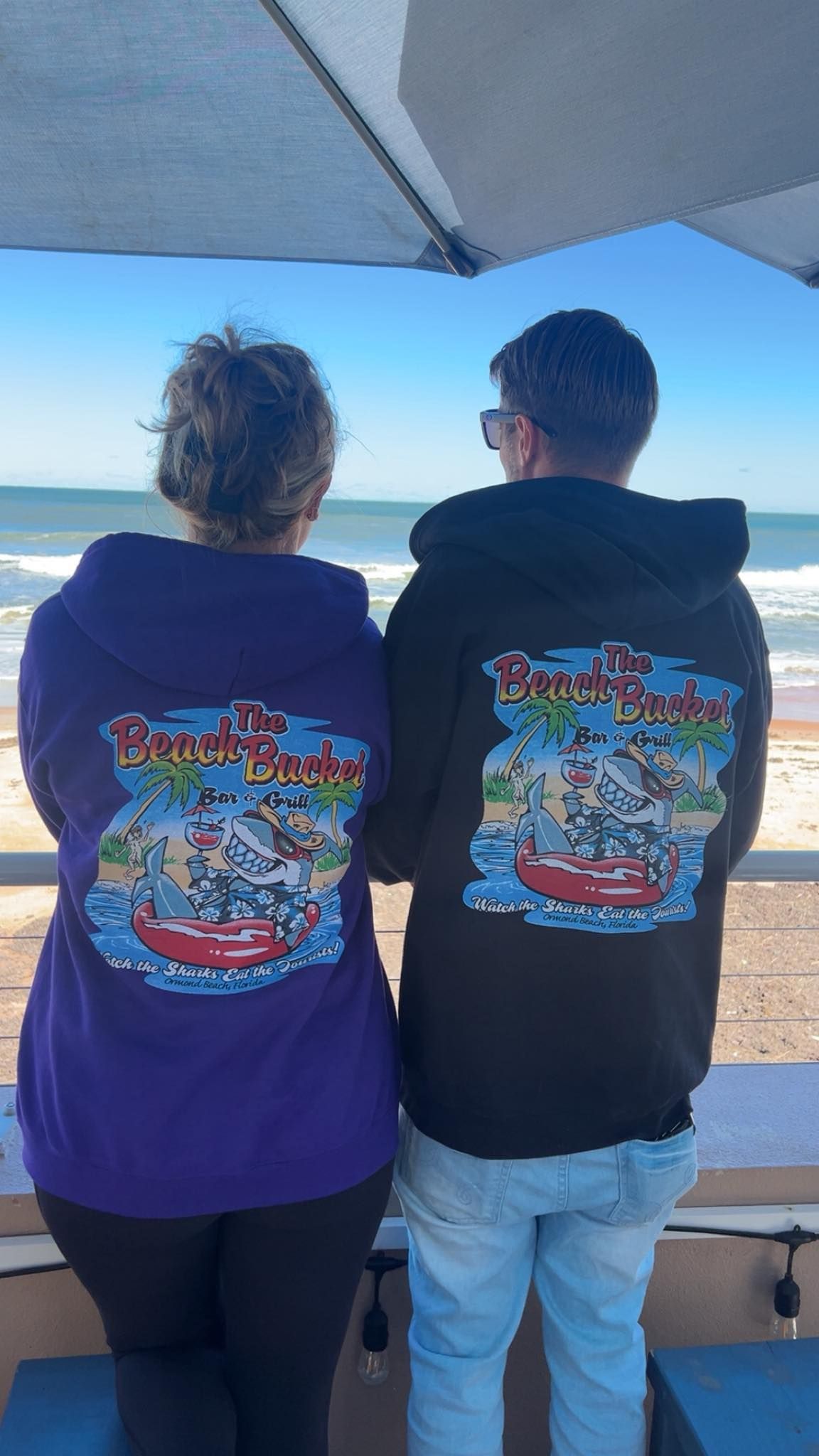 Two people facing a beach wearing hoodies with The Beach Bucket logo on the back.