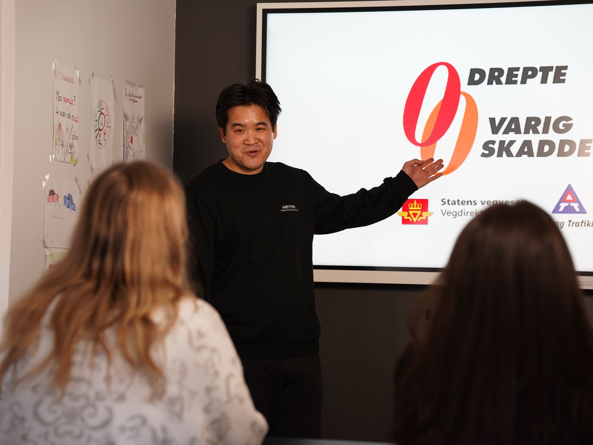 Foredragsholder presenterer ved siden av et projisert lysbilde med tekst og logoer «Dreptie va rid skadade»