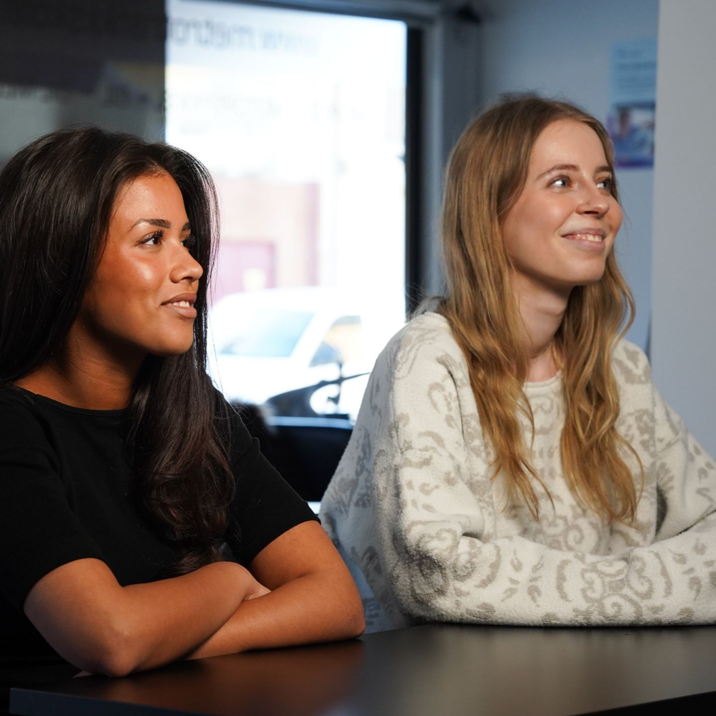 To kvinner sitter ved et bord, smiler og lytter i et lyst møterom.
