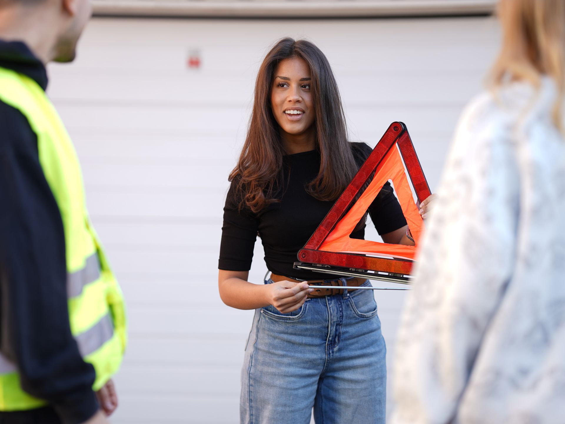 Kvinne som holder en rød varseltrekant, står mellom to personer i en garasje.