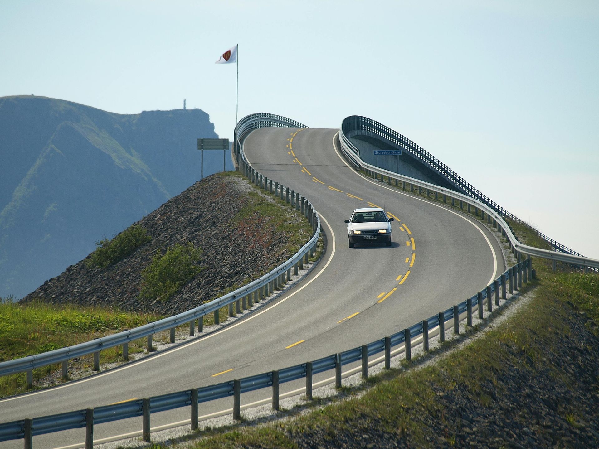 Bil på en svingete fjellvei med autovern og et flagg mot en klar himmel