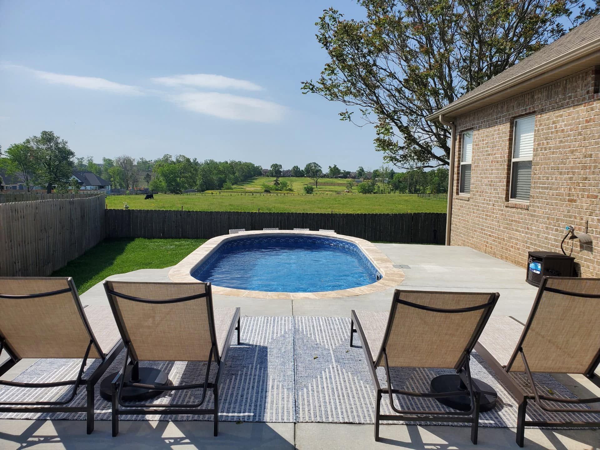 Backyard patio with four lounge chairs facing a blue swimming pool and open green field beyond