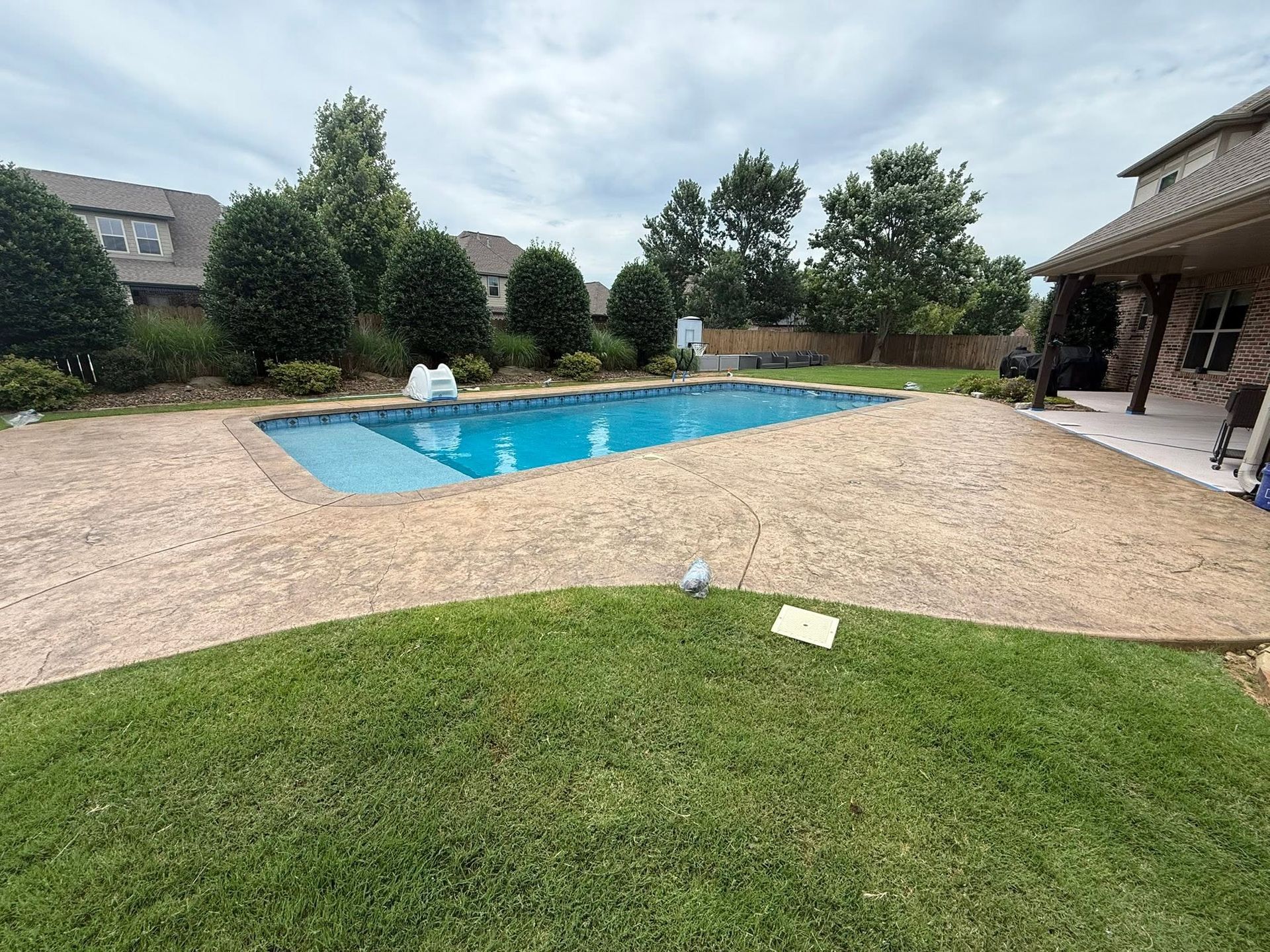 Backyard swimming pool with blue water, beige gravel patio, green lawn, and trees under a cloudy sky