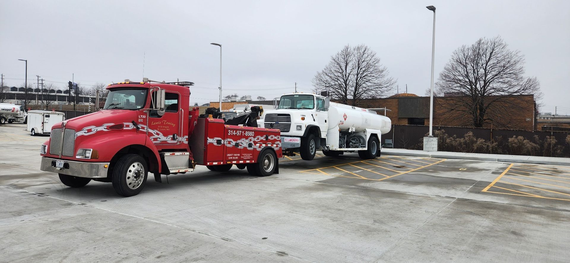 A red tow truck is towing a white truck in a parking lot.