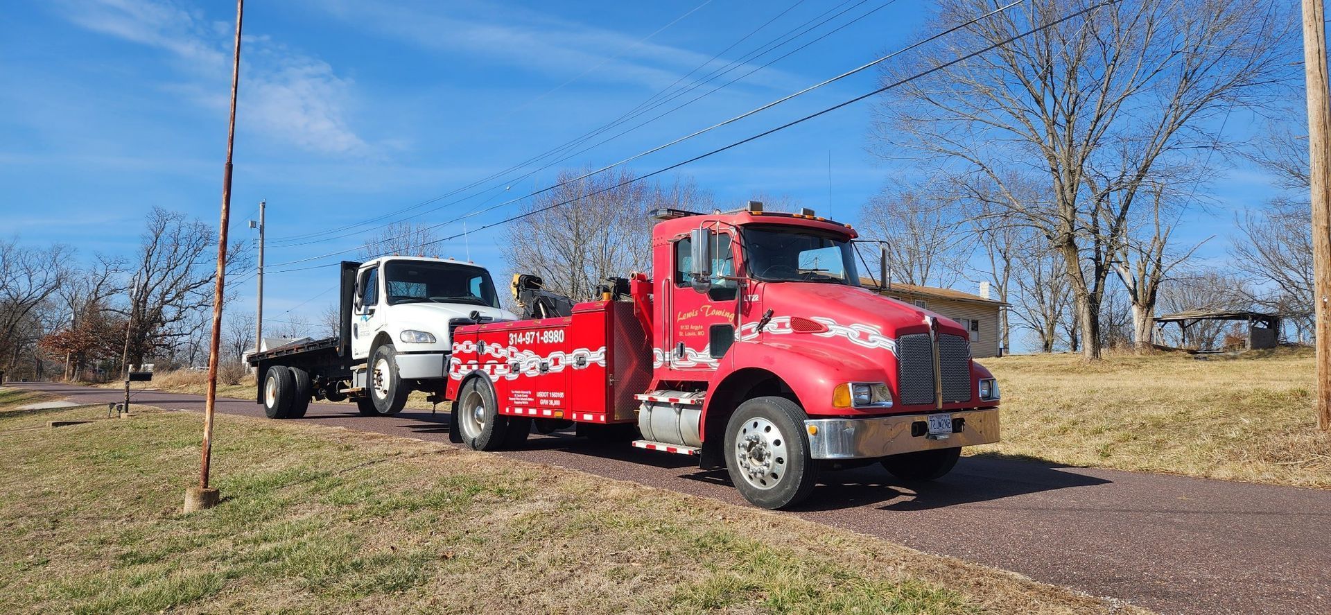 A red tow truck is towing a white truck on a dirt road.