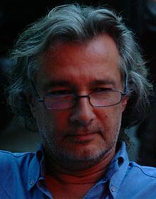 Gros plan sur un homme aux cheveux gris et portant des lunettes, vêtu d’une chemise bleue et regardant l’objectif.