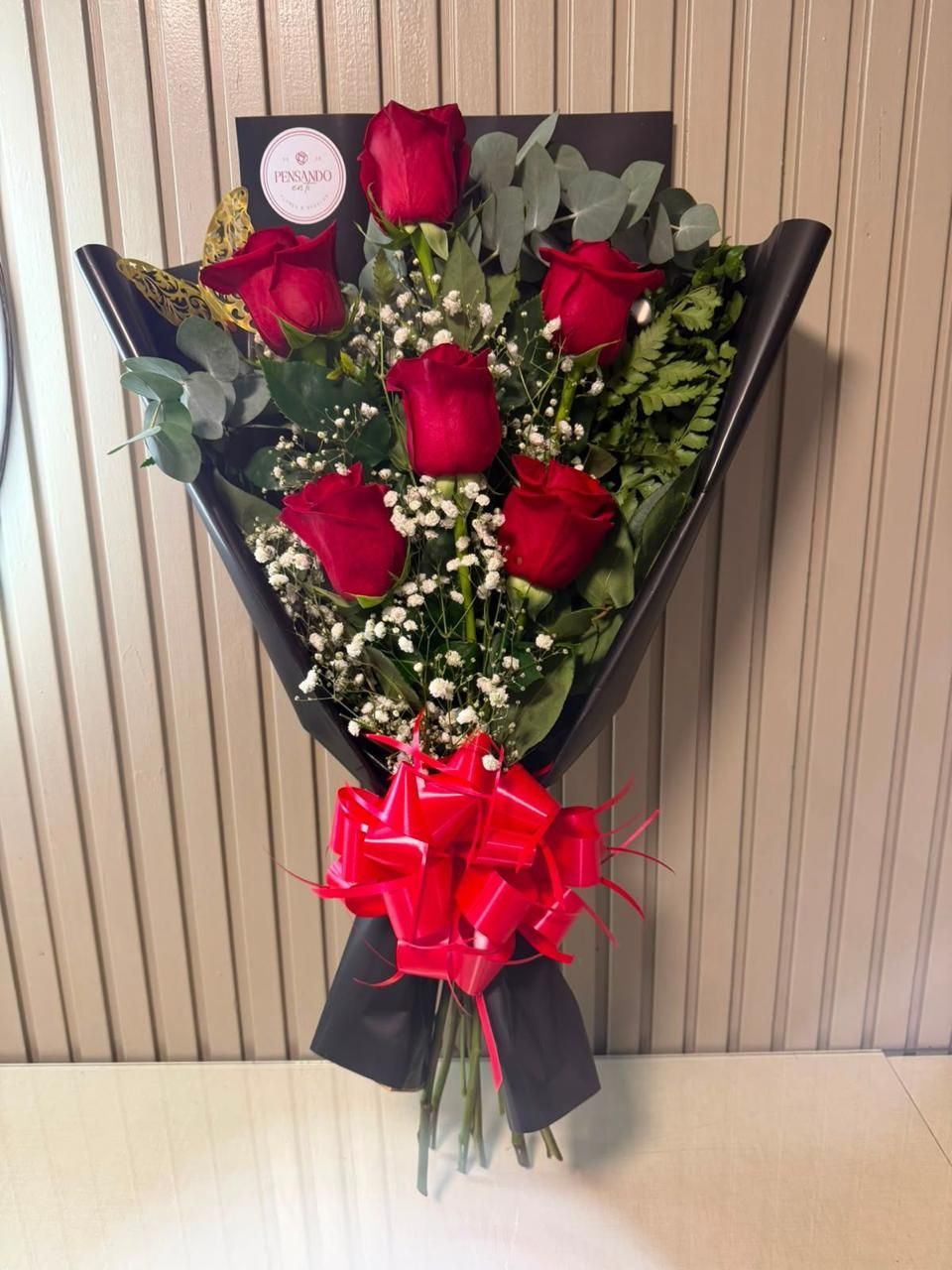Ramo de seis rosas rojas con follaje, envuelto en papel negro y atado con una cinta roja.