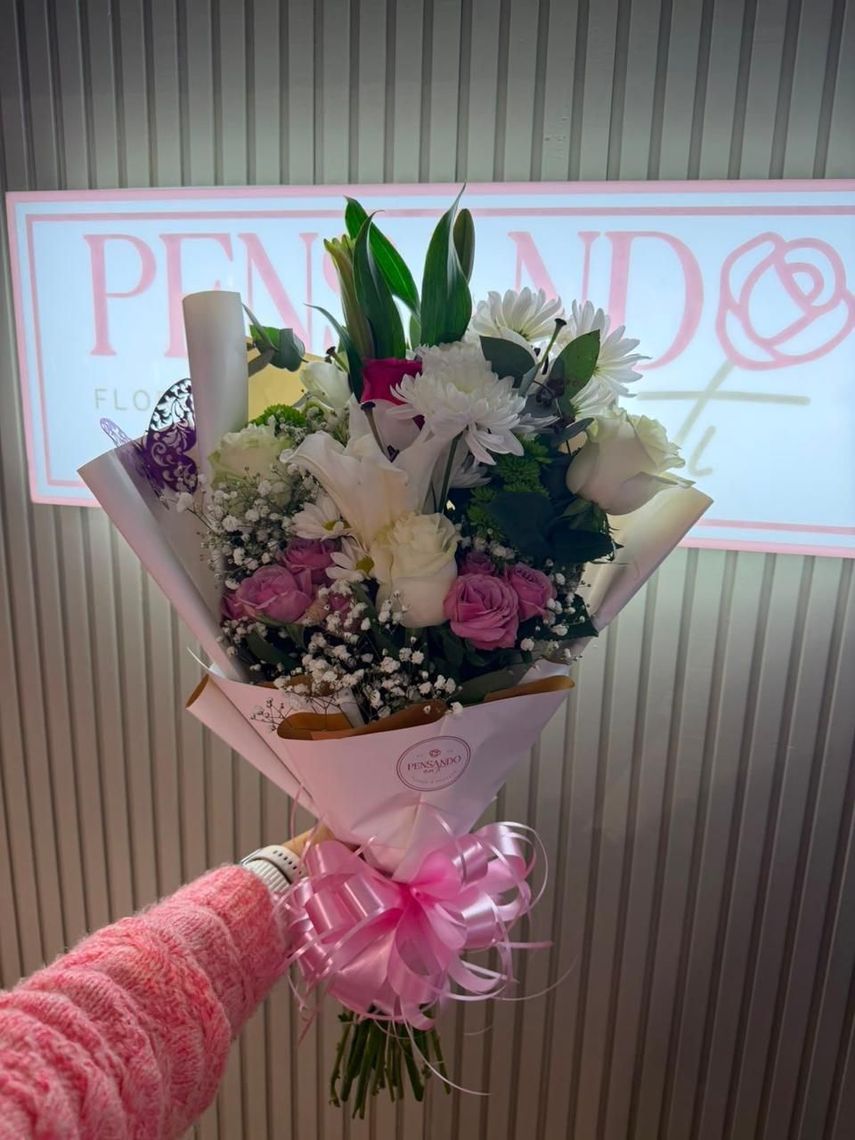 Ramo de flores blancas y moradas envuelto en papel rosa, sostenido frente a un letrero de una tienda.