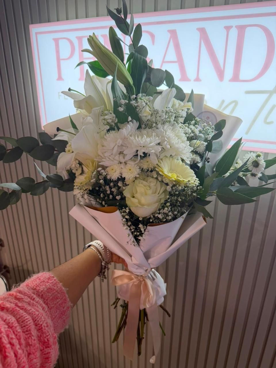 Ramo blanco con rosas y follaje envuelto en papel rosa pálido, sostenido frente al letrero de una tienda.