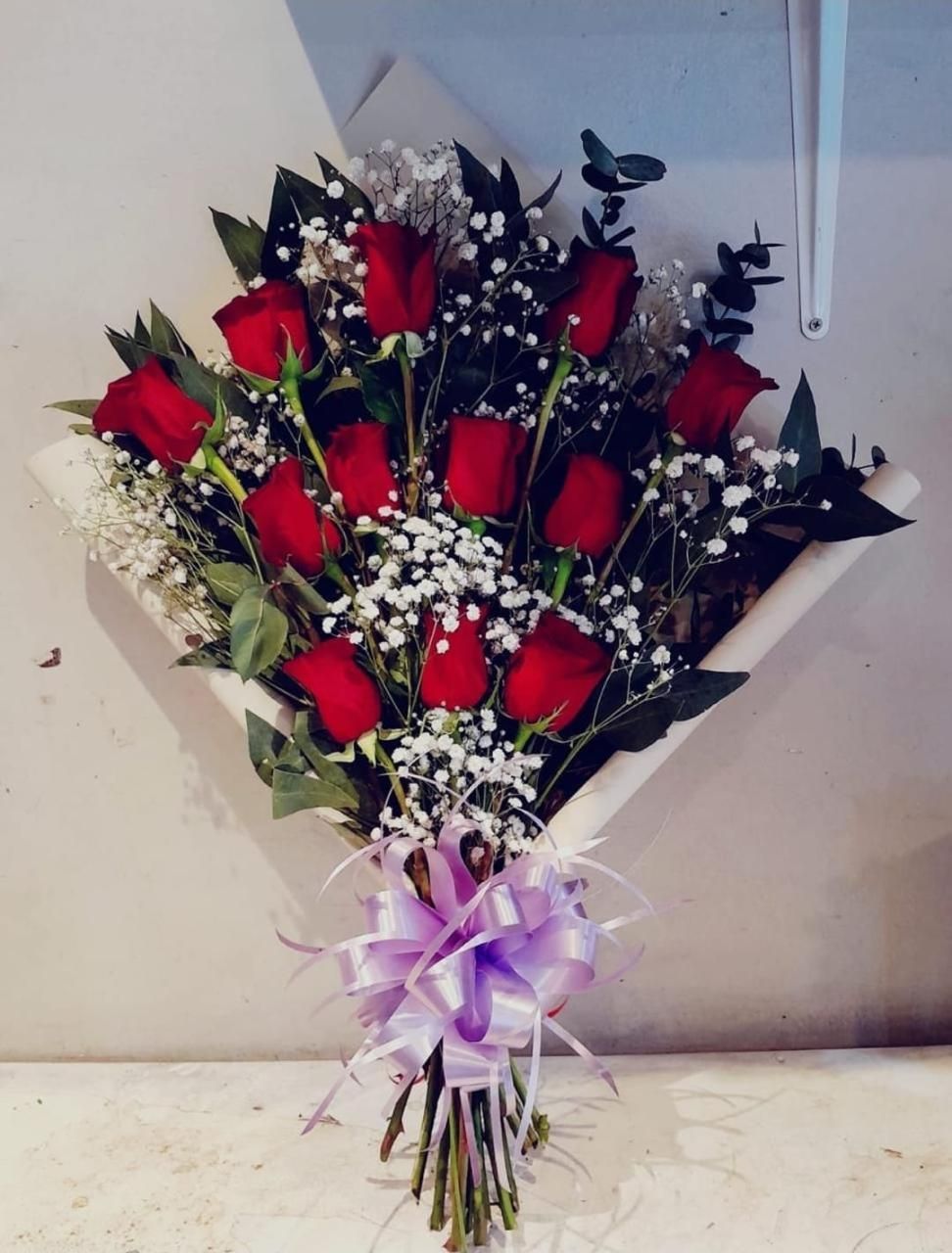 Ramo de rosas rojas con paniculata blanca y cinta morada, envuelto en papel negro sobre una superficie clara.