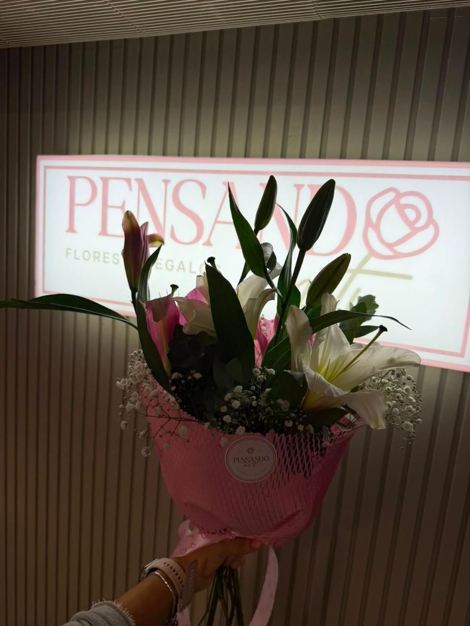Ramo de flores rosas sostenido frente a un letrero iluminado de “Pensa” en el interior.