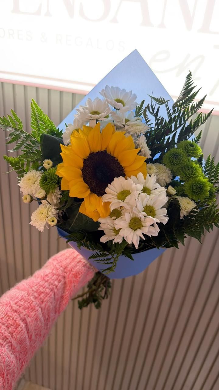 Ramo de mano con girasoles, margaritas blancas y follaje, envuelto en papel azul.