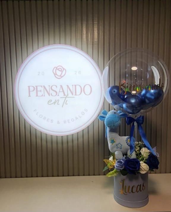 Arreglo de regalo con globo transparente, cintas azules, flores y un letrero iluminado