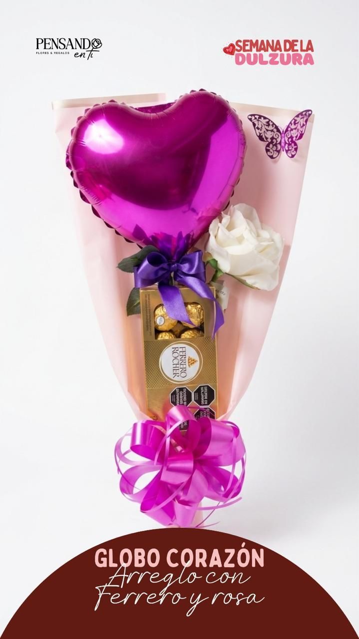 Cesta de regalo rosa con forma de corazón, adornada con globos, flores y chocolate sobre un fondo blanco.