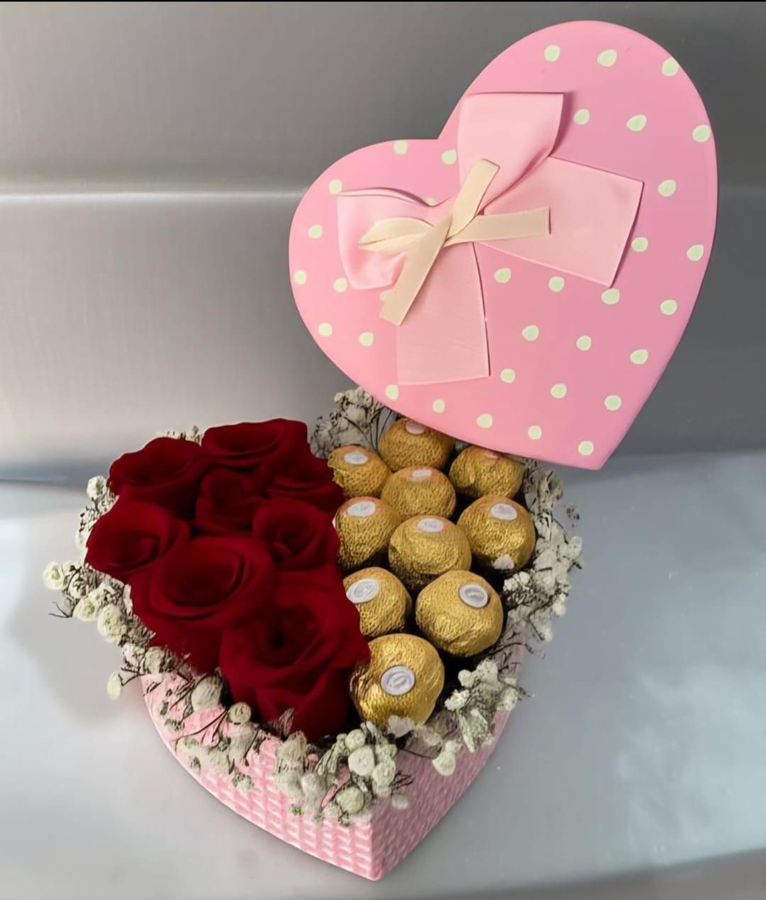 Caja de regalo en forma de corazón con rosas rojas, bombones dorados y una tapa con lazo rosa sobre una superficie gris.