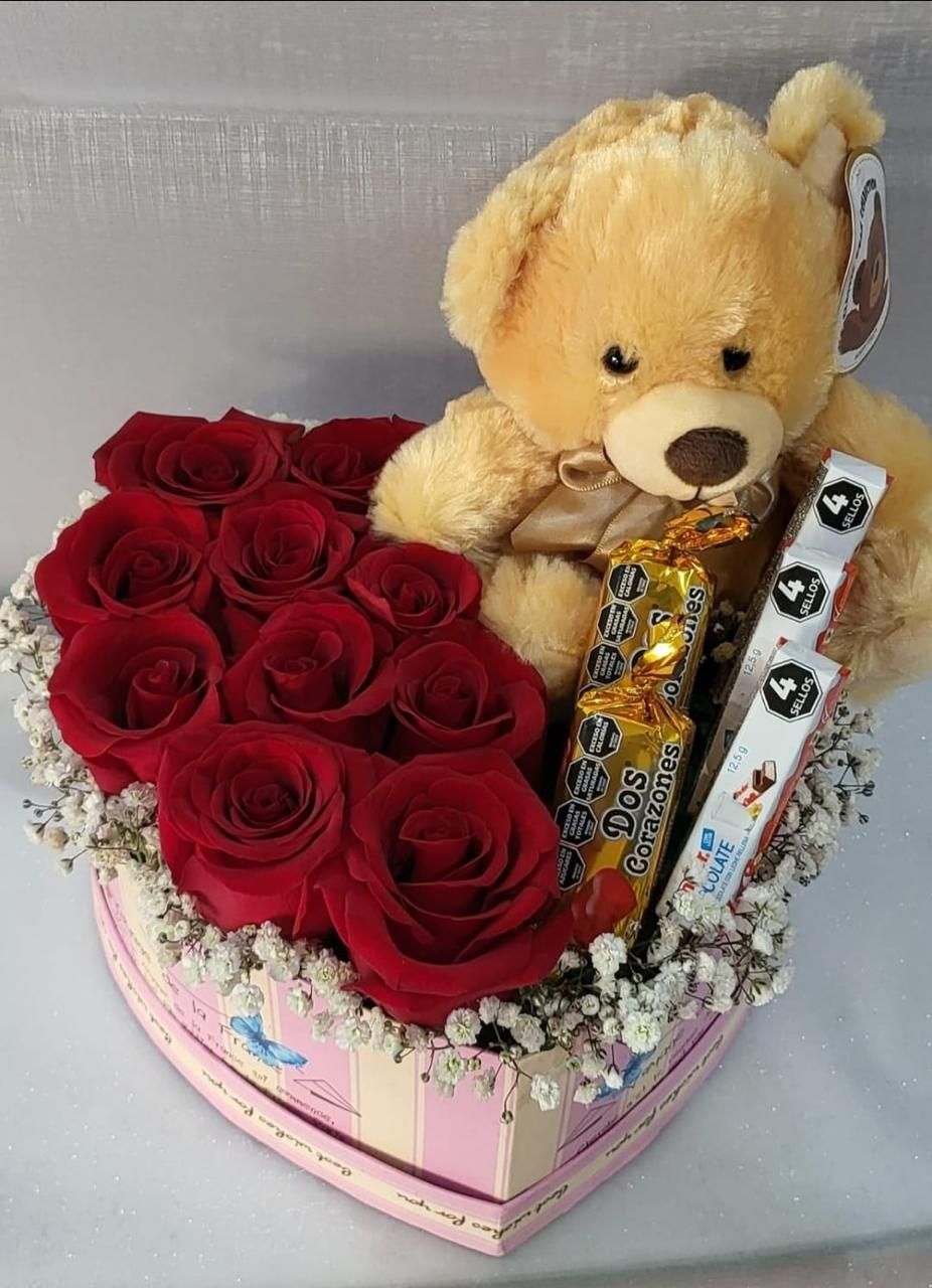 Osito de peluche con rosas rojas y bombones envueltos en una caja rosa con forma de corazón.