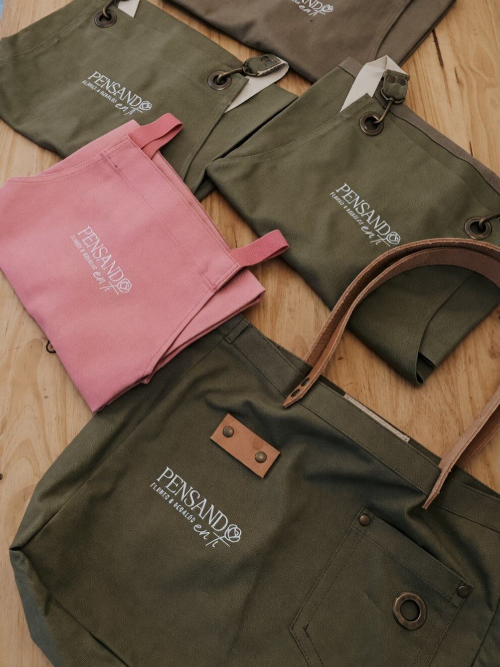 Bolsas de lona variadas en verde oliva y rosa, dispuestas sobre un suelo de madera.
