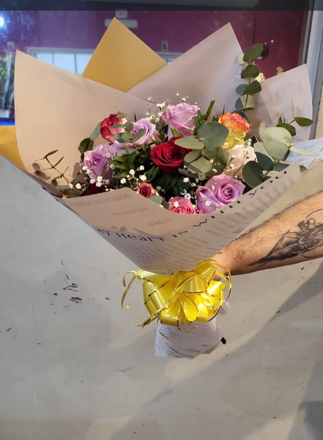 Ramo de flores rosas y blancas envuelto en papel color crema con una cinta amarilla, sostenido sobre un suelo de cemento.