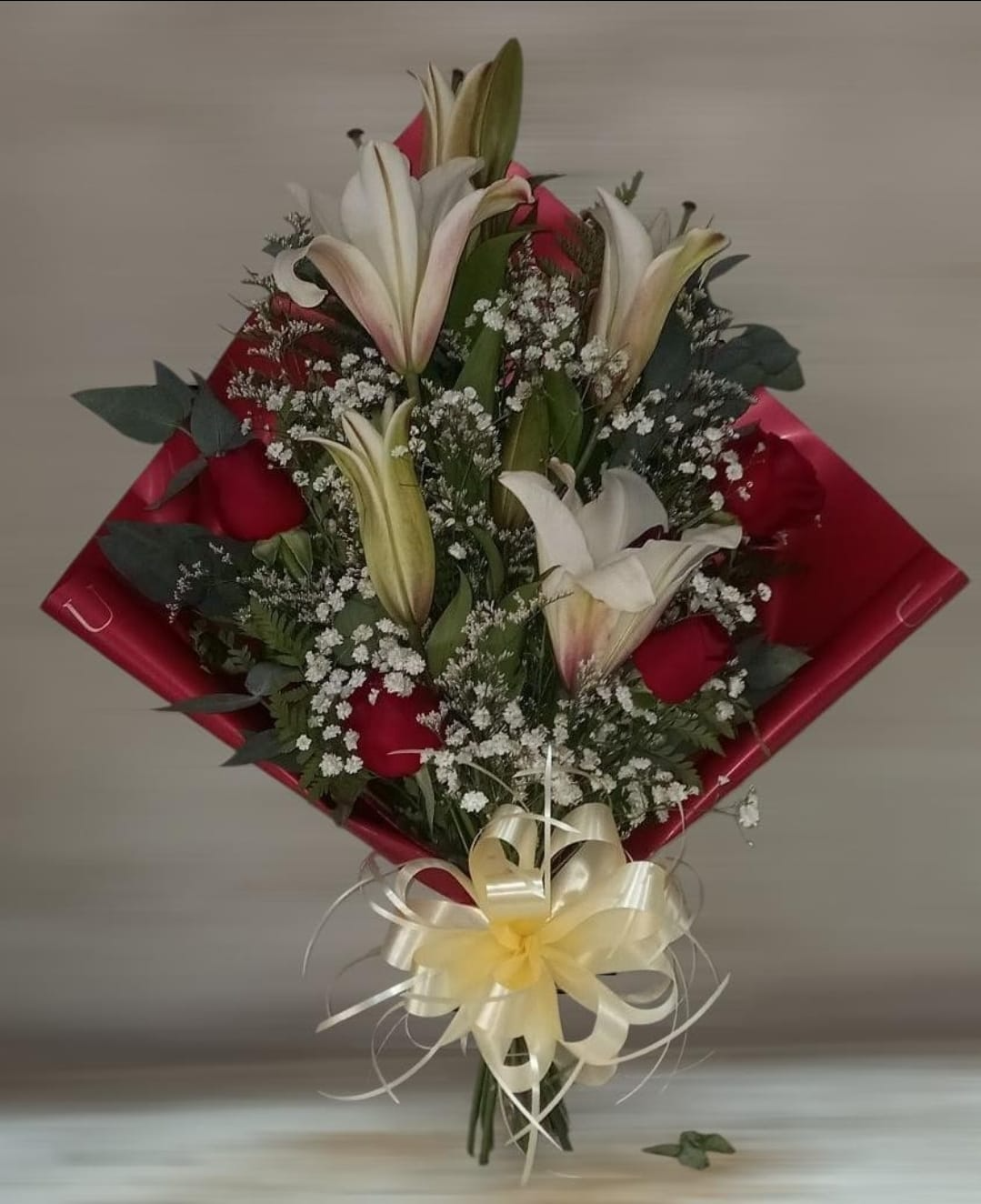 Ramo de lirios blancos, rosas rojas y paniculata envuelto en papel rojo con una cinta blanca.