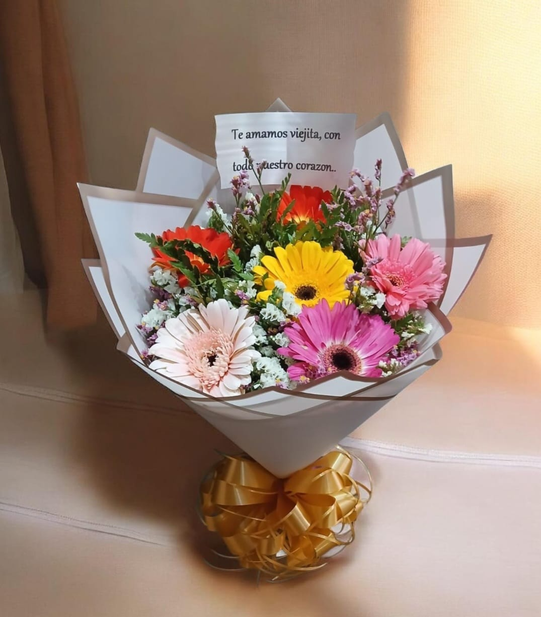 Ramo de flores rosas, amarillas y blancas envuelto en papel con una cinta dorada sobre una mesa.