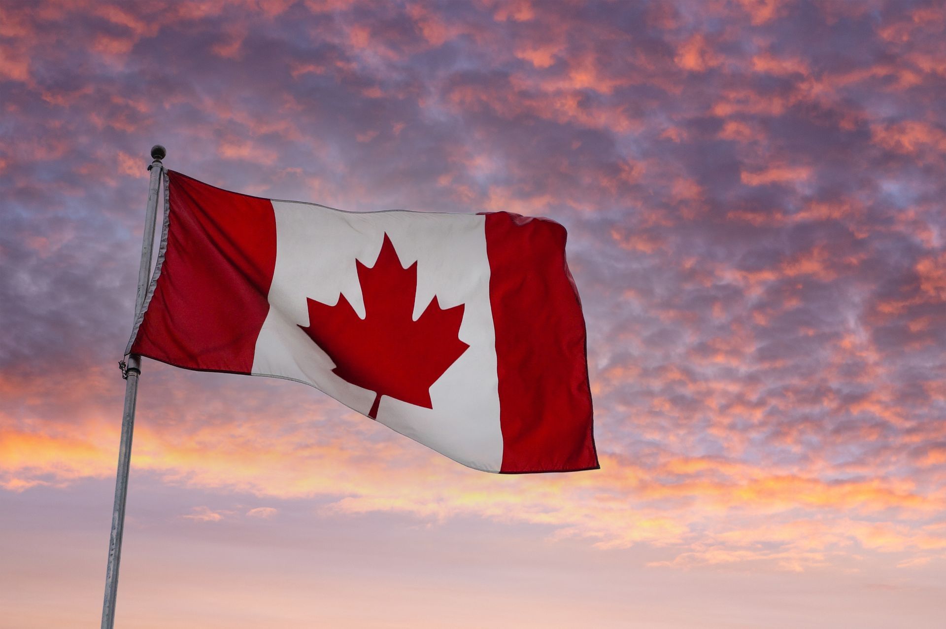 The Canadian flag waves on a flagpole against a backdrop of a colorful, cloudy sunrise sky.