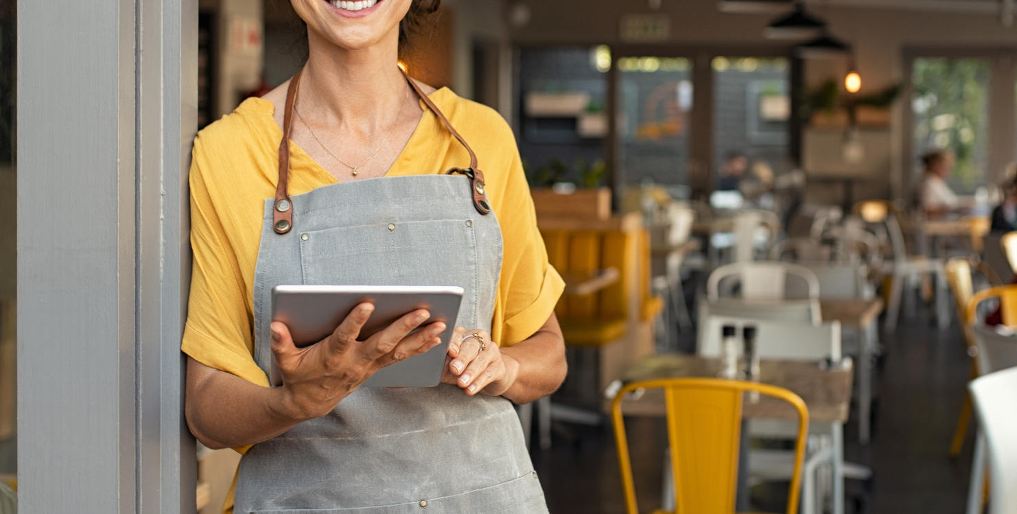 Smiling café worker in yellow shirt and gray apron holding a tablet inside a coffee shop