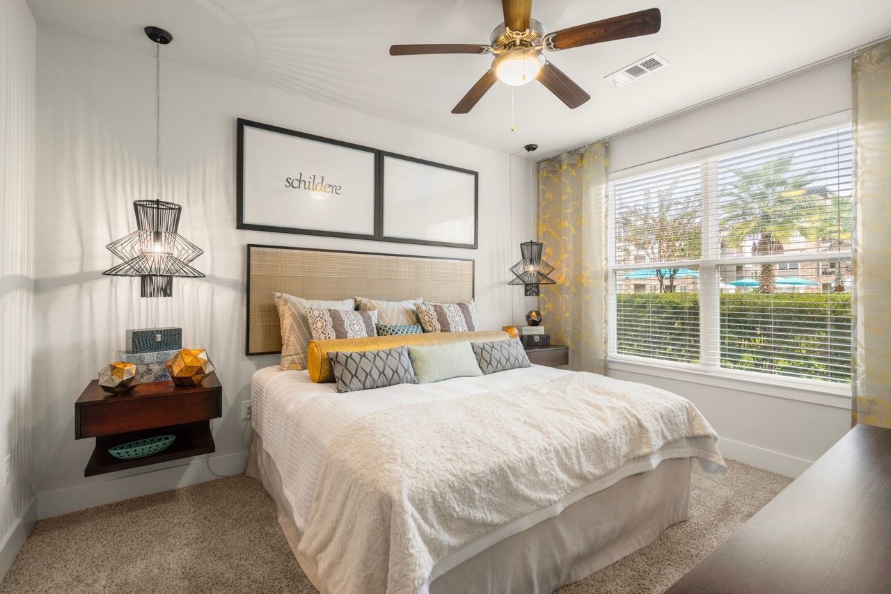 Bedroom in an apartment with a large bed, two nightstands, ceiling fan, and a window with blinds.