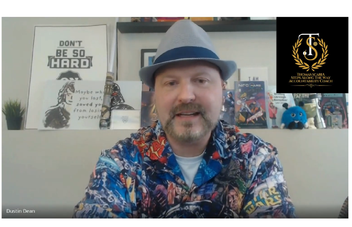 Man in blue hat and floral shirt speaking on webcam, with posters and bookshelves behind him.