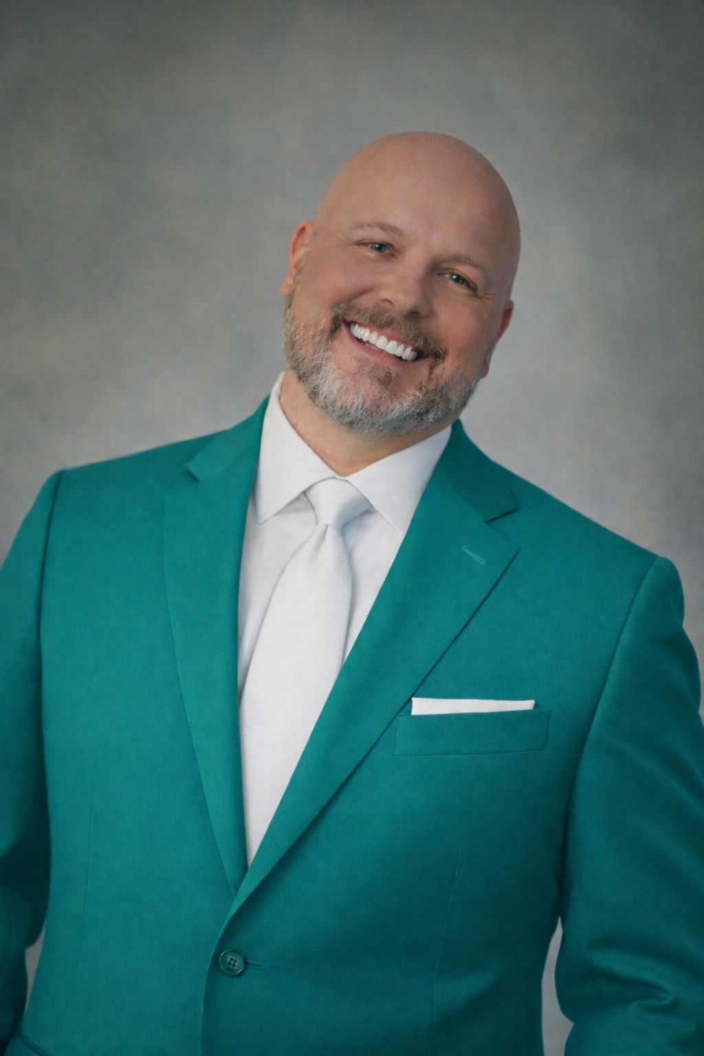 Smiling man in a teal suit and white tie against a gray studio backdrop