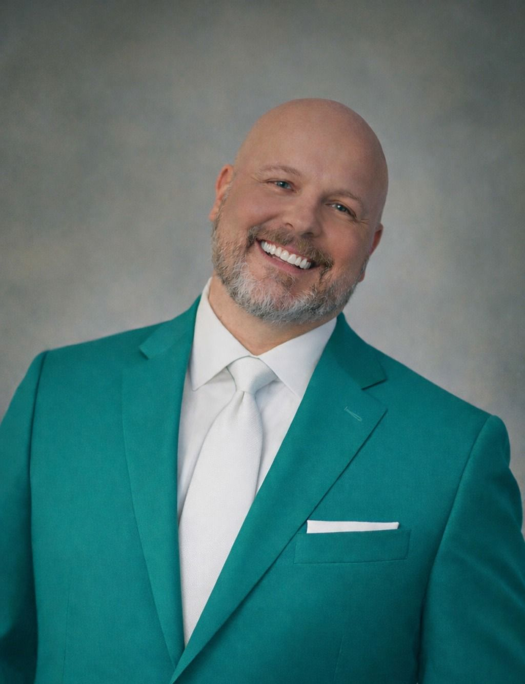 Smiling bald man in a teal suit and white tie against a gray studio backdrop