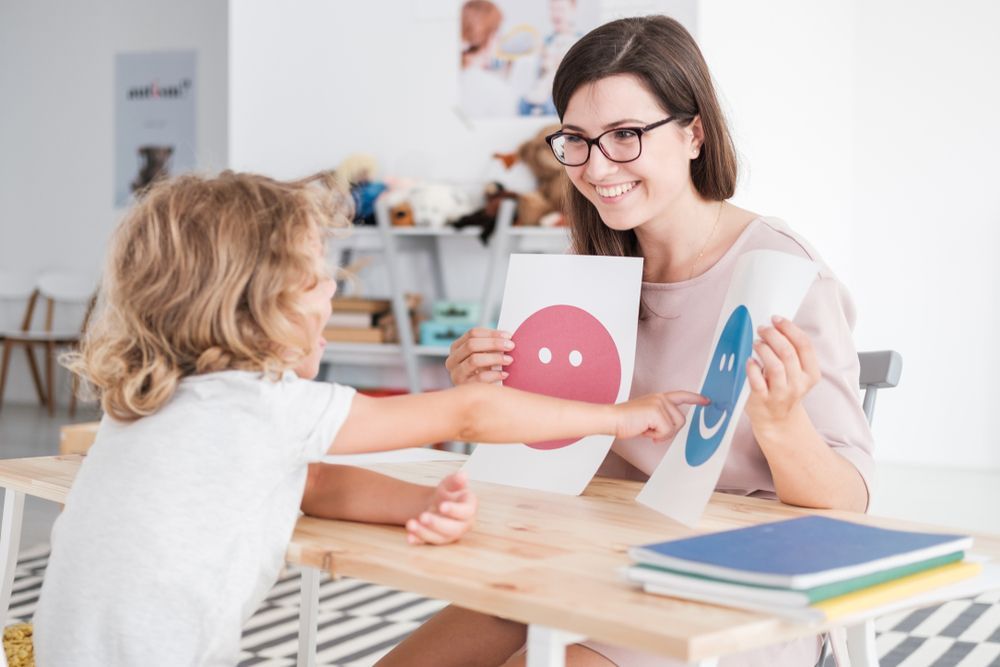 Woman showing colorful face cards to a child at a table in a bright classroom