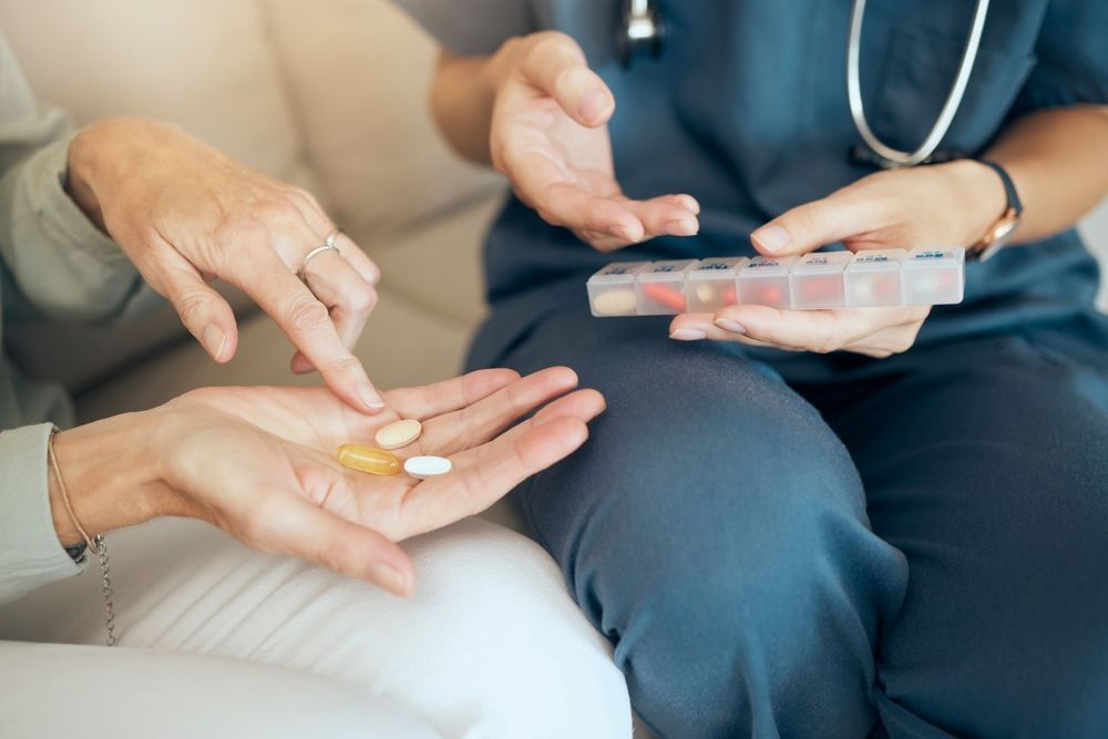 Healthcare worker handing pills to a patient seated on a couch, with a prescription bottle in hand