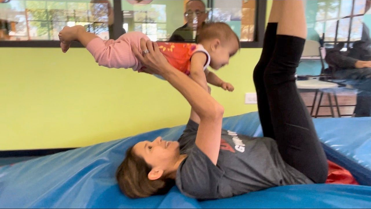 Woman lying down lifting a baby in the air. The baby is wearing an orange shirt and pink pants, lying with arms out.