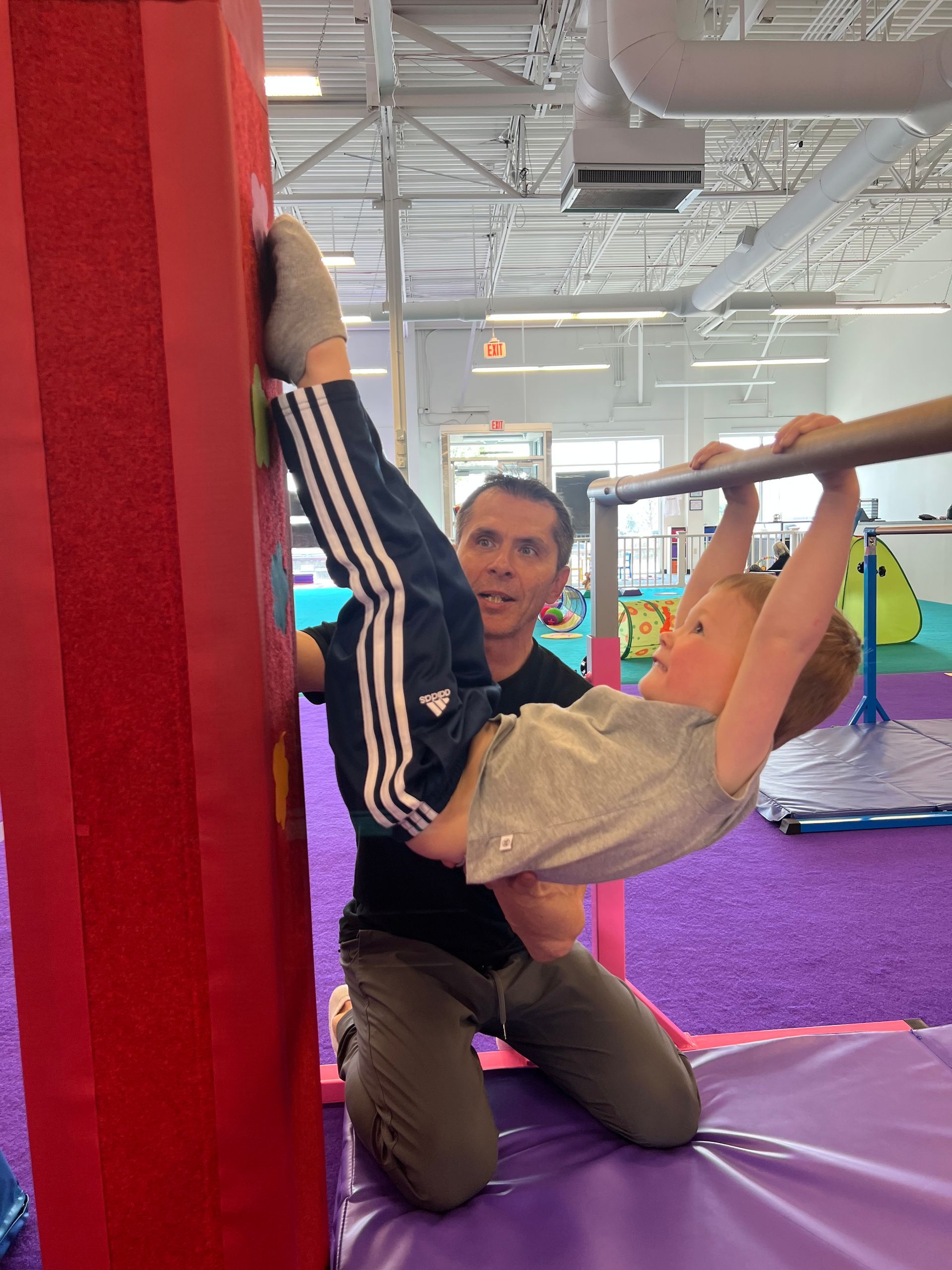 A girl doing a backbend on a blue mat, assisted by an instructor in a yellow shirt. Other children watch.