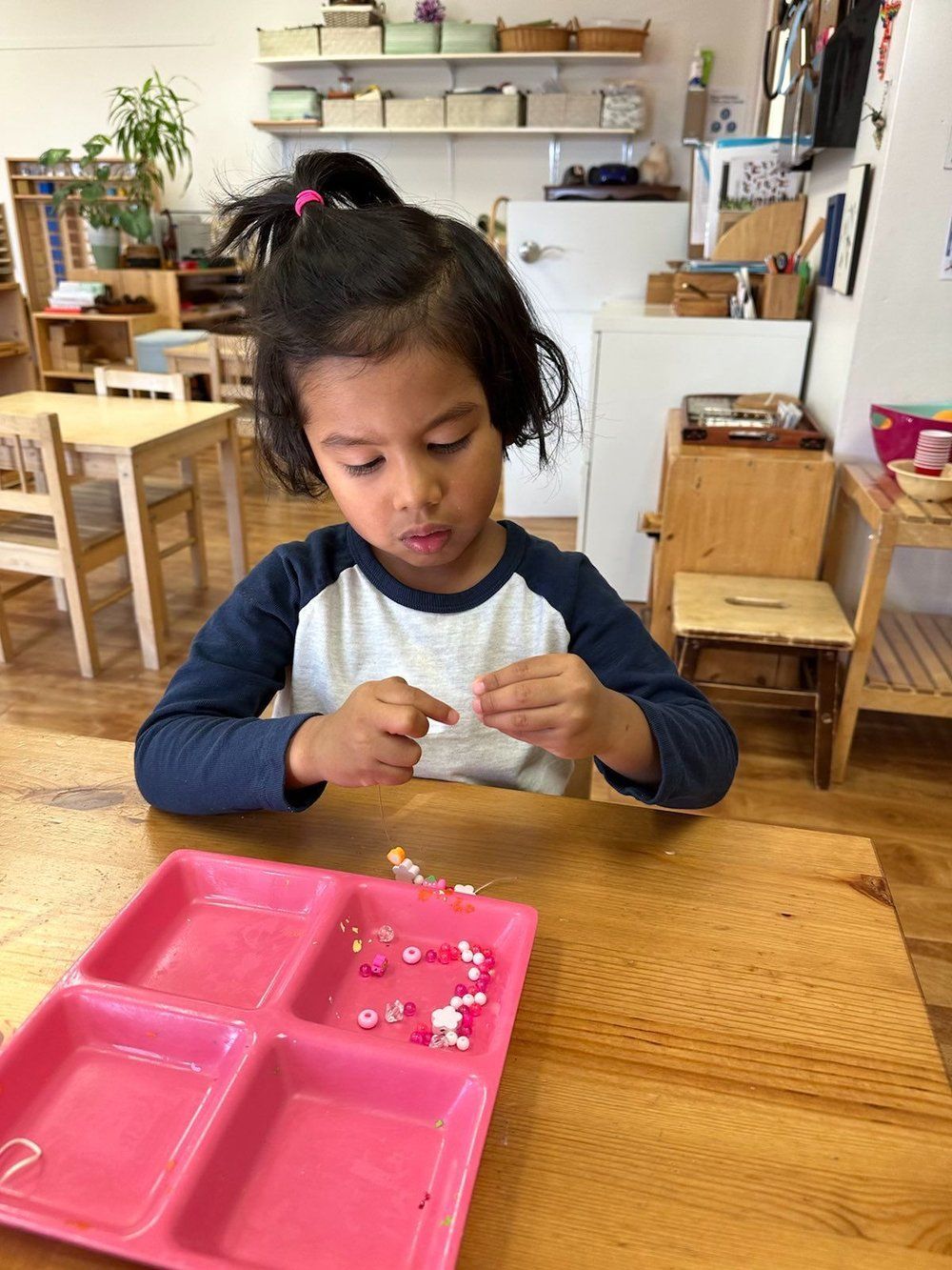 A little girl is sitting at a table playing with a pink tray.
