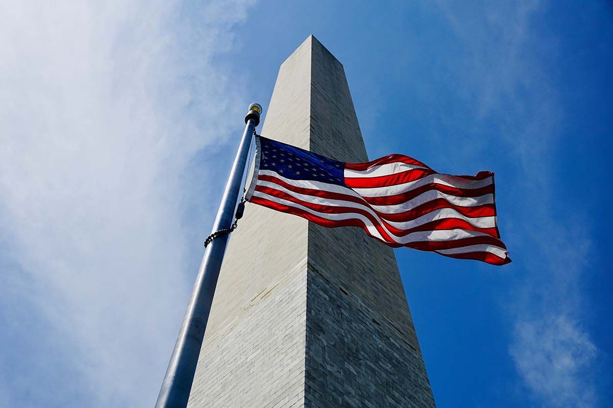 American flag waving on a pole beside the Washington Monument against a blue sky