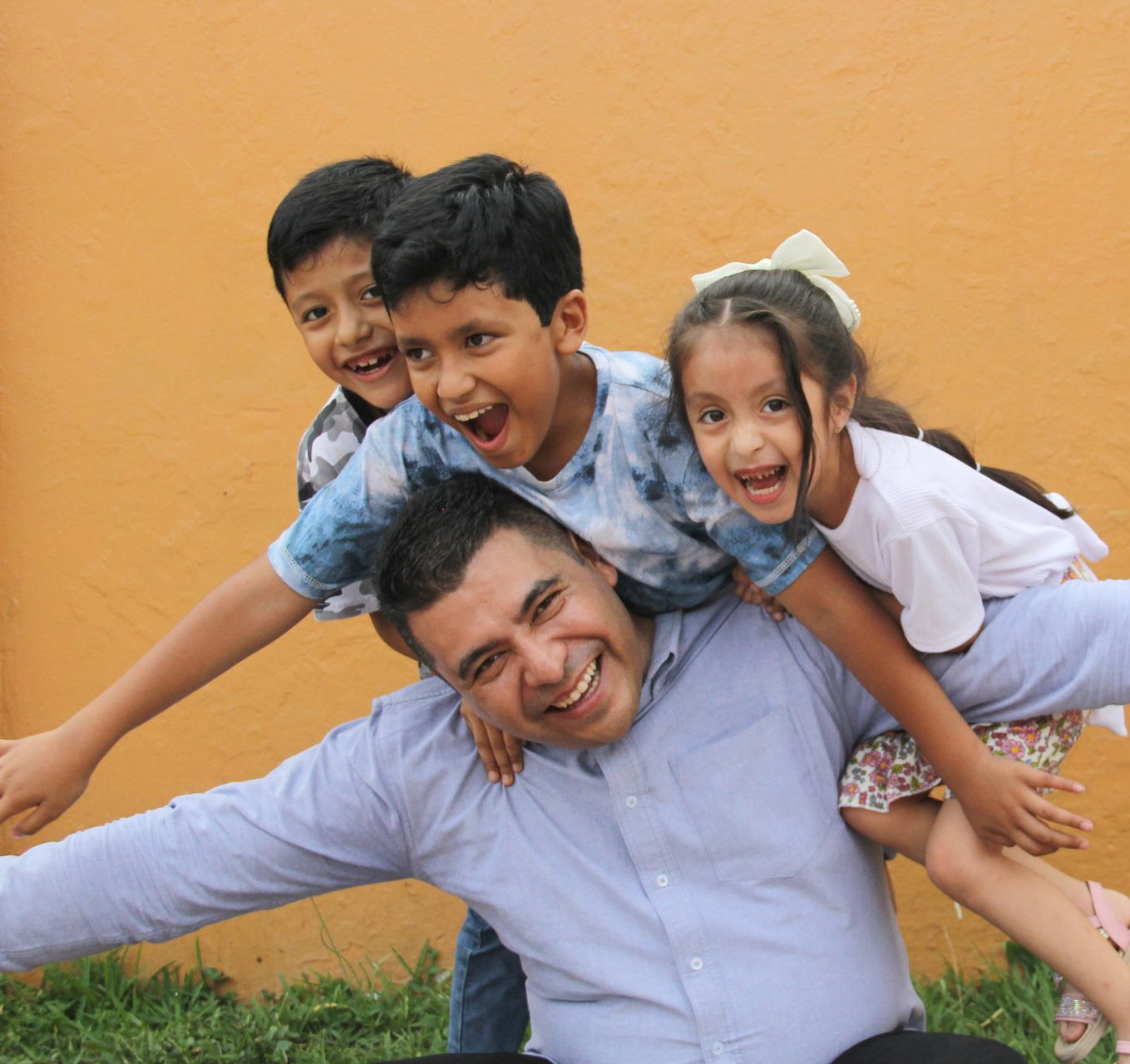 Smiling man giving two laughing children a piggyback ride against an orange wall