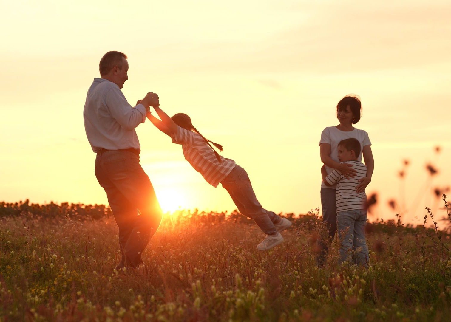 Family playing in a field at sunset, with one adult swinging a child and another standing nearby.