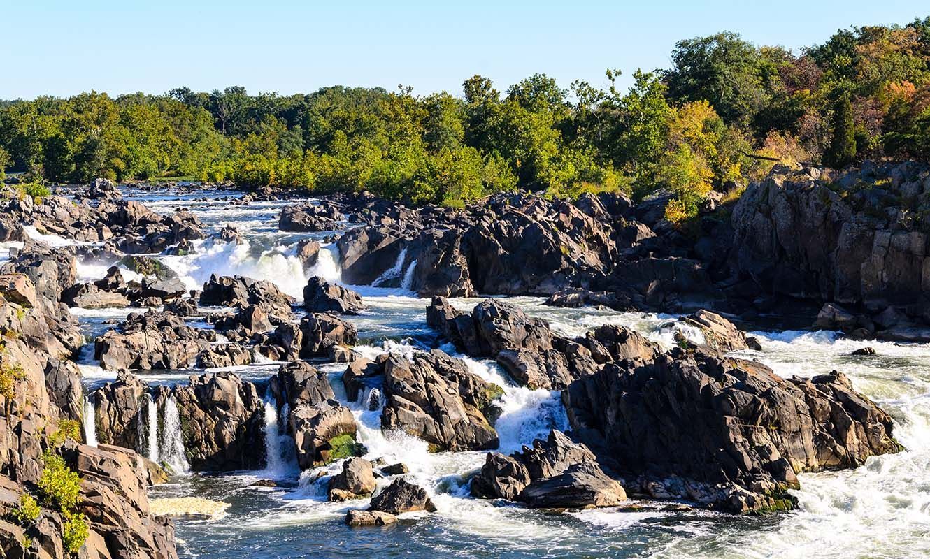 Rugged waterfall cascading over dark rocks with white water and green trees under a clear sky