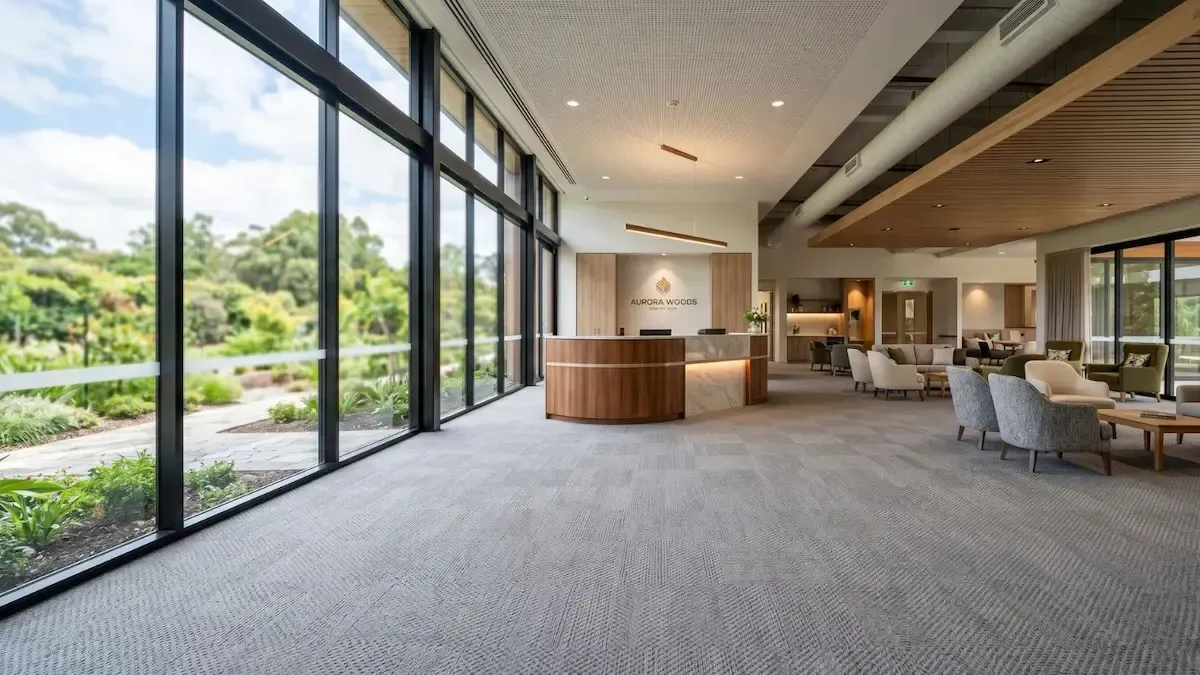 Modern hotel lobby with floor-to-ceiling windows, carpeted seating area, and wood reception desk overlooking greenery