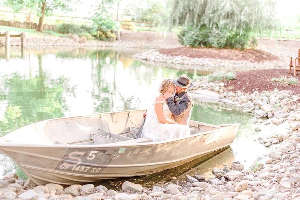 Couple in wedding attire kissing in a boat on a pond, rocks in foreground, weeping willow in background.