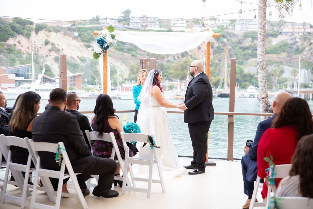 Wedding ceremony by the water with bride, groom, and seated guests under white chairs and floral arch.