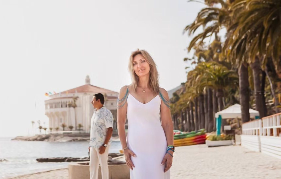 Woman in white dress smiles on beach, man in background, palm trees, building.